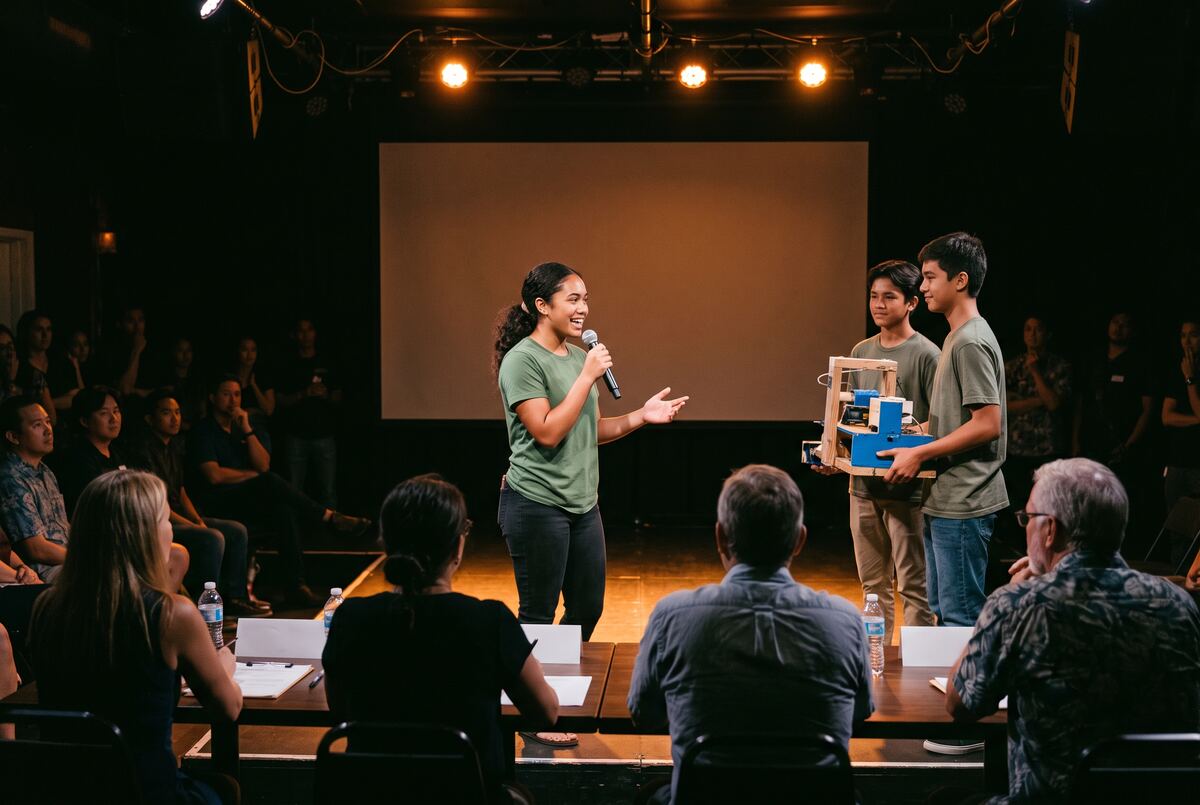 A student pitches with a microphone while teammates hold their prototype in front of judges.